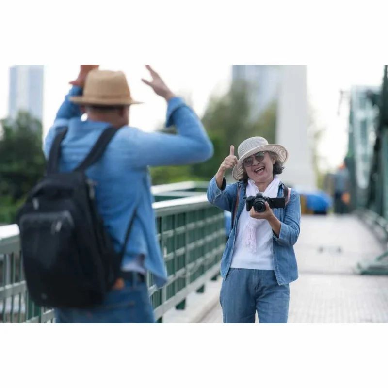 Ältere Frau mit Kamera und Daumen hoch, Mann im Hintergrund. Auf einer Brücke.