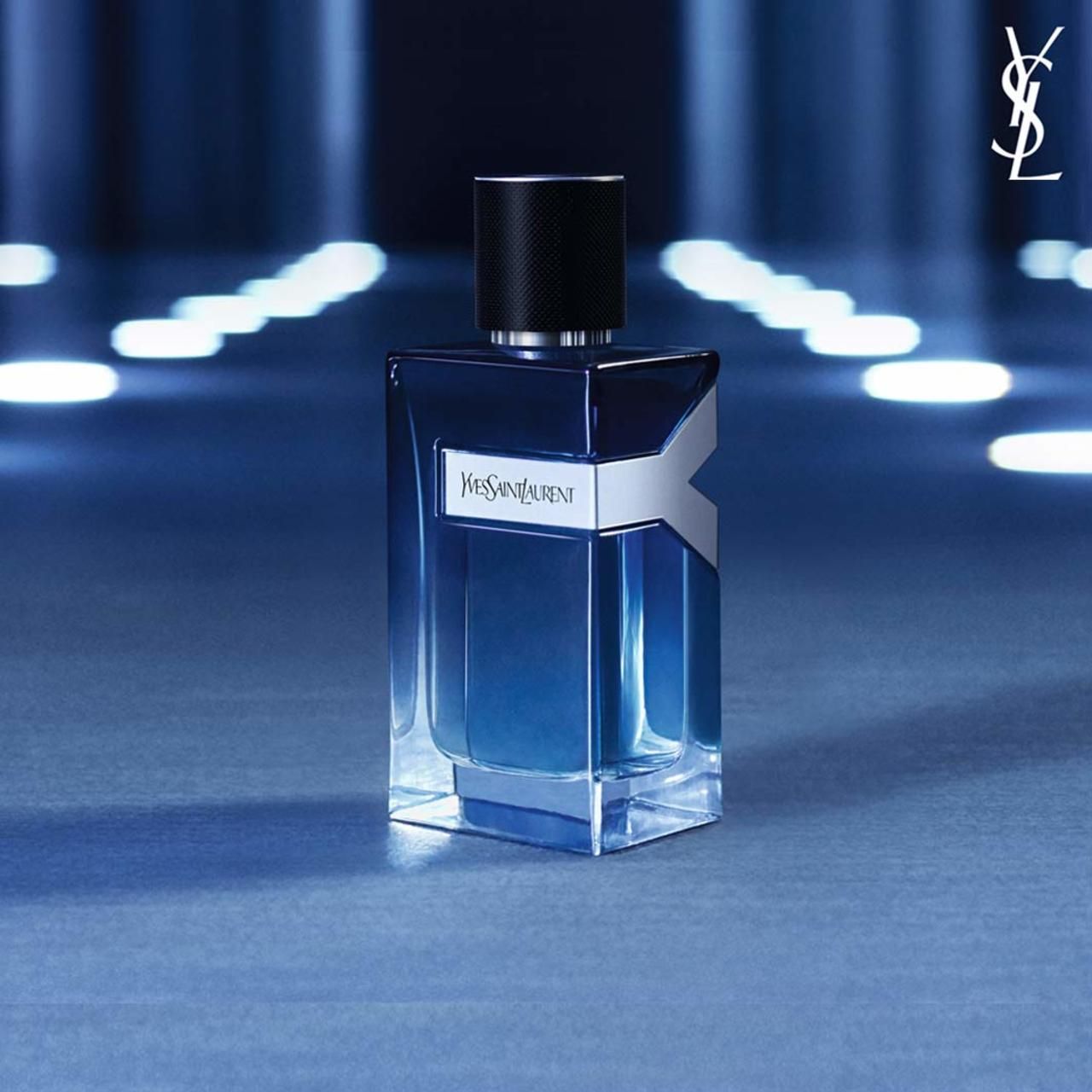Blauer Parfümflakon mit schwarzem Deckel. Silbernes Logo. YSL-Schriftzug. Auf blauem Hintergrund.