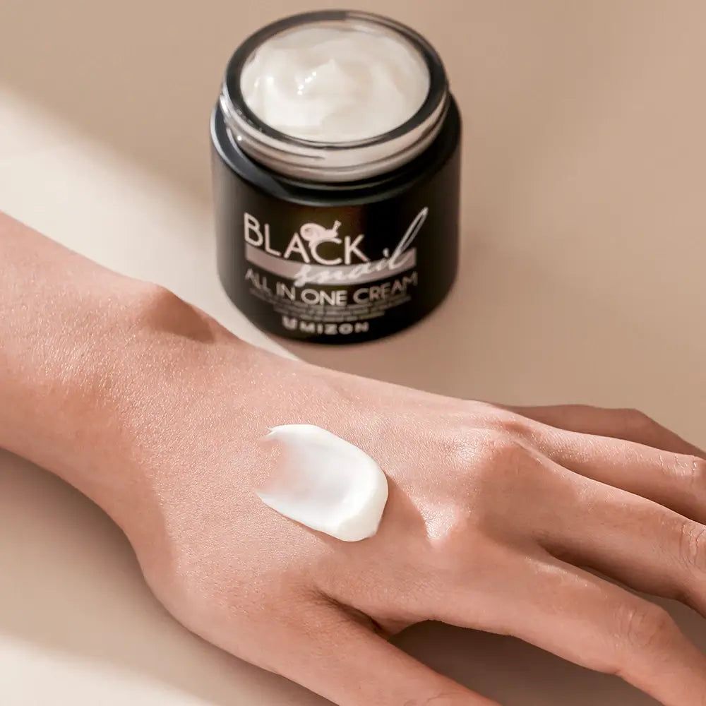 Offener schwarzer Tiegel mit Creme. Creme auf Hand aufgetragen. Aufschrift: BLACK snail ALL IN ONE CREAM.