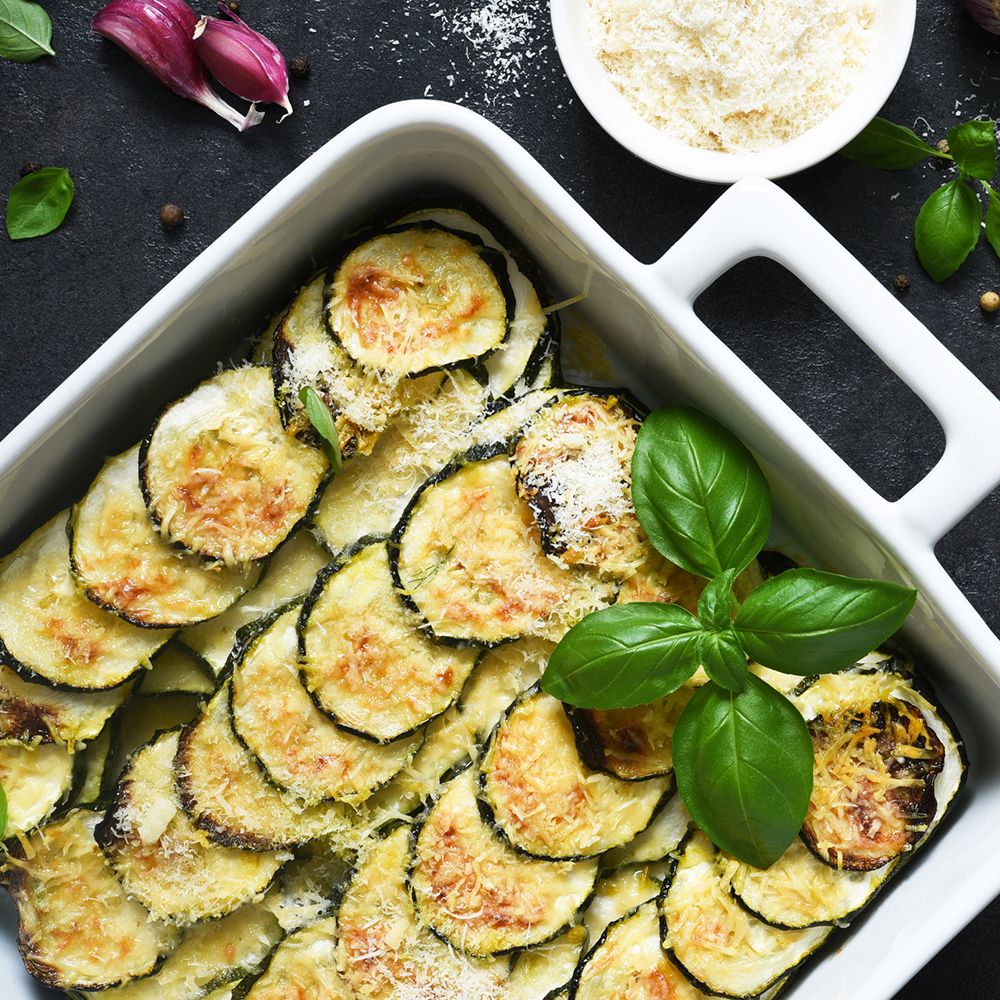 Auflaufgericht mit Zucchinischeiben, Käse und Basilikumblättern. Weißer Auflaufform auf dunklem Untergrund.