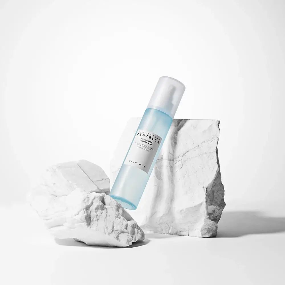 Blau-transparente Sprühflasche auf weißen Steinen. Aufschrift: MADAGASCAR CENTELLA, HYALU-CICA Cloudy Mist. SKIN1004.