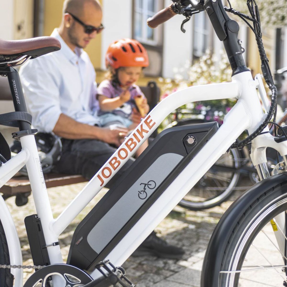 Weißes E-Bike mit Akku, JOBOBIKE-Logo. Mann und Kind sitzen auf Bank im Hintergrund.