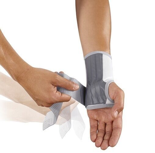 Handgelenkbandage, grau-weiß, wird mit Klettverschluss befestigt. Eine Hand legt die Bandage an, die andere fixiert den Verschluss.