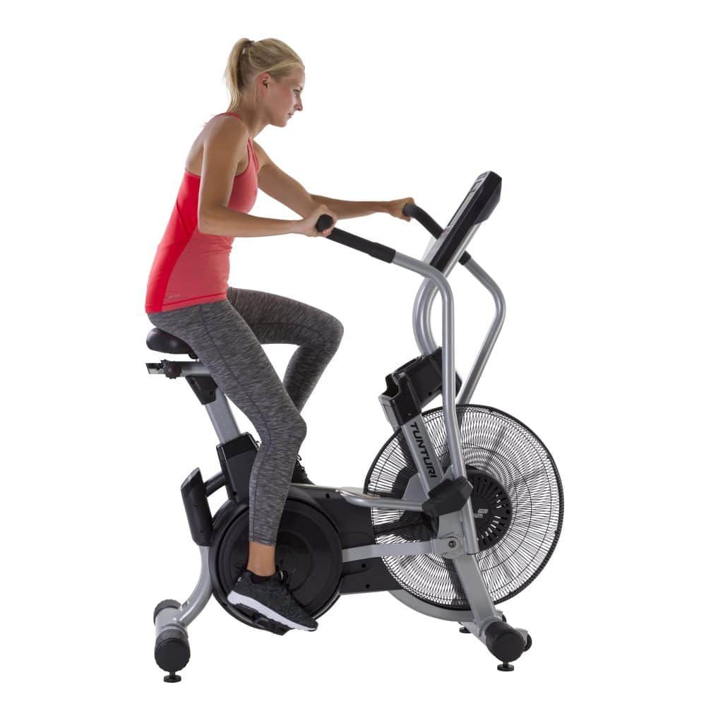 Frau auf Airbike. Rotes Top, graue Hose. Hält Lenker. Ventilator sichtbar. Tunturi-Logo.