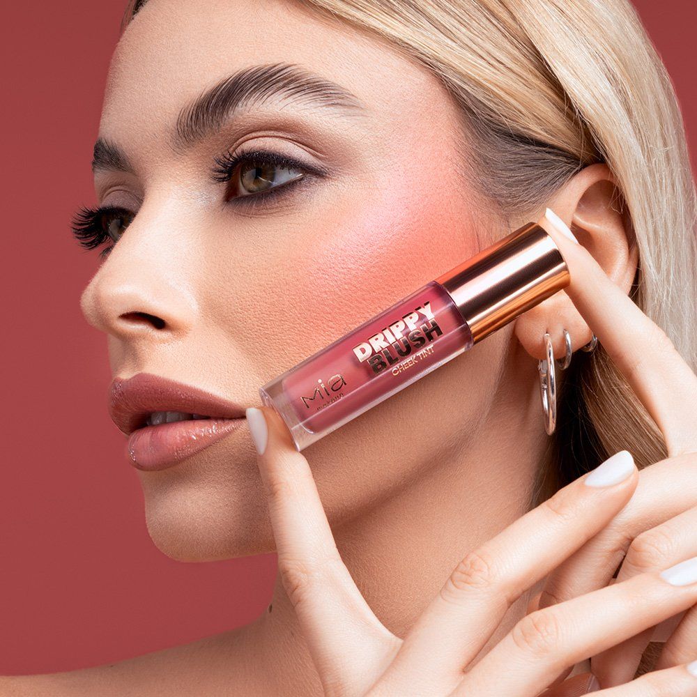 Una donna tiene in mano un tubetto di Mia Make-up Drippy Blush. Il tubetto ha un tappo color oro rosa e contiene una tonalità rosa. Il testo "Drippy Blush" è visibile sul tubetto. La donna ha applicato il blush sulle guance.