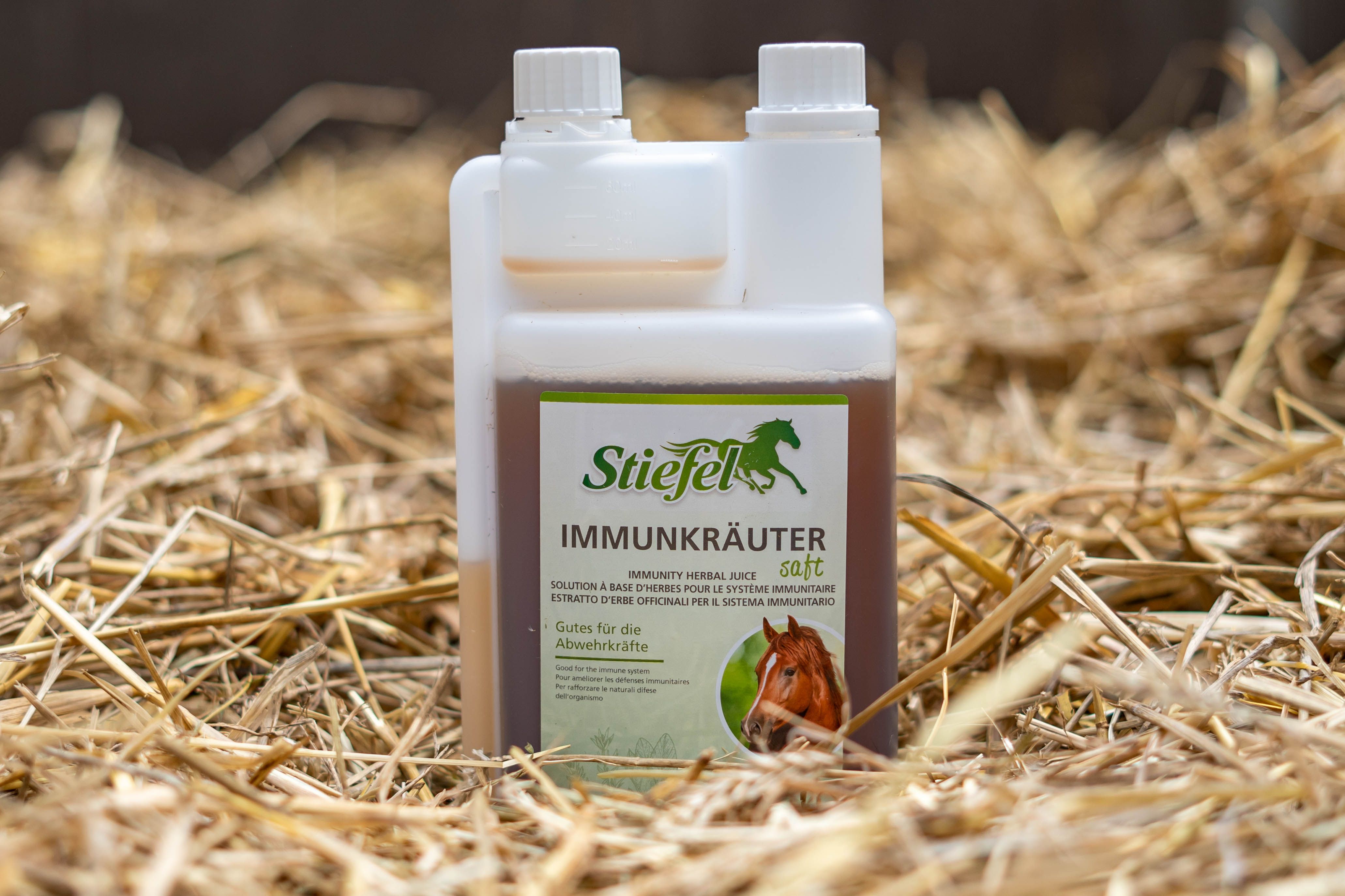 Rechteckige Flasche mit brauner Flüssigkeit, weißem Deckel und Etikett. Liegt auf Stroh. Etikett mit Produktnamen und Logo.