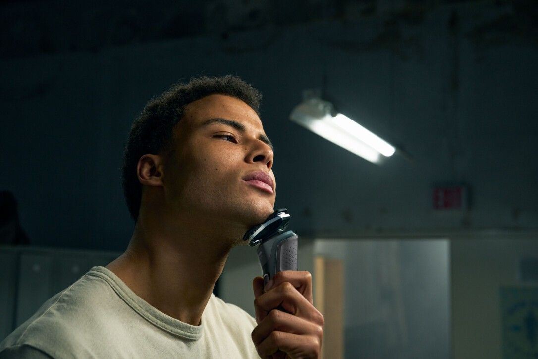 Mann rasiert sich mit einem elektrischen Rasierer. Helles T-Shirt. Beleuchtung von oben.