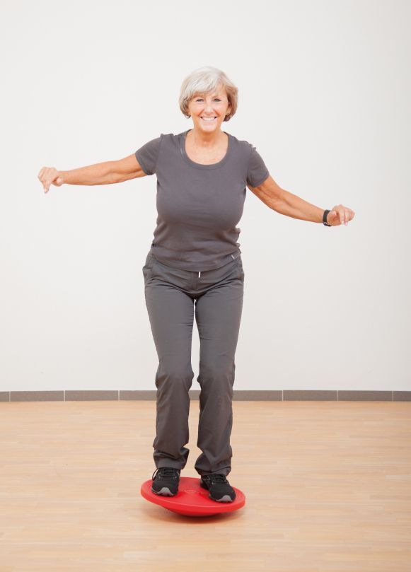 Ältere Frau steht auf rotem Therapiekreisel. Arme seitlich ausgestreckt. Sportkleidung.