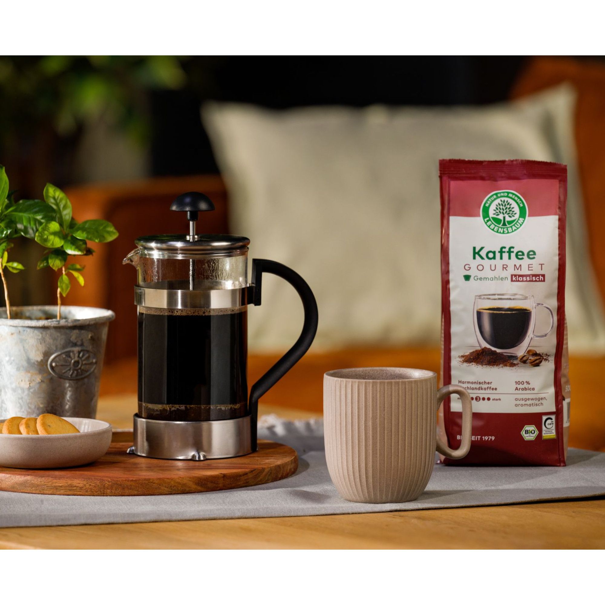 Glas-Kaffeepresse mit Edelstahl-Akzenten und einer Packung "Kaffee Gourmet" daneben.