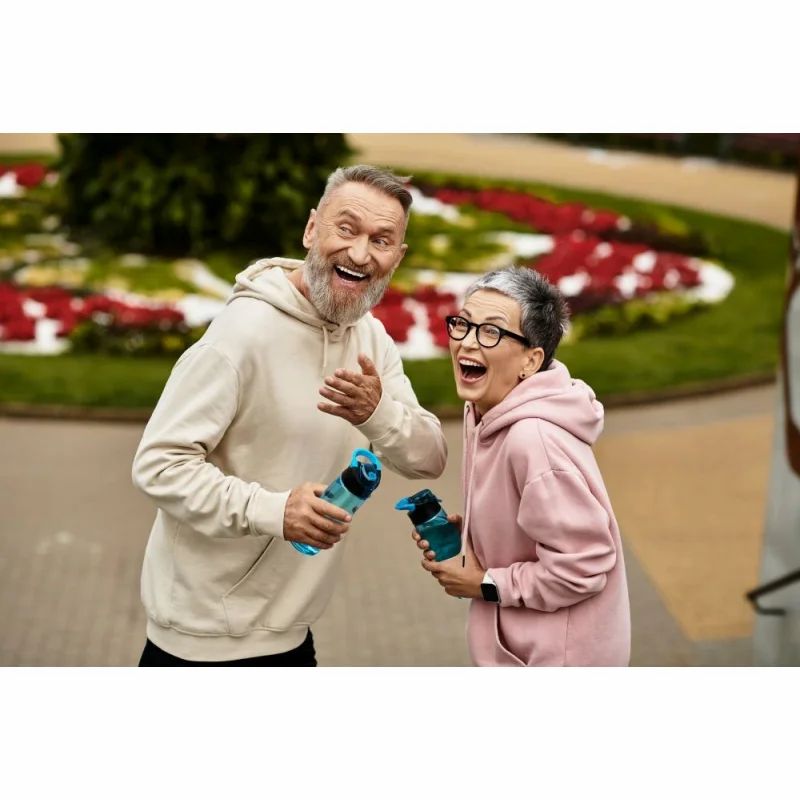 Zwei ältere Menschen lachen und halten blaue Trinkflaschen. Beide tragen Kapuzenpullover.
