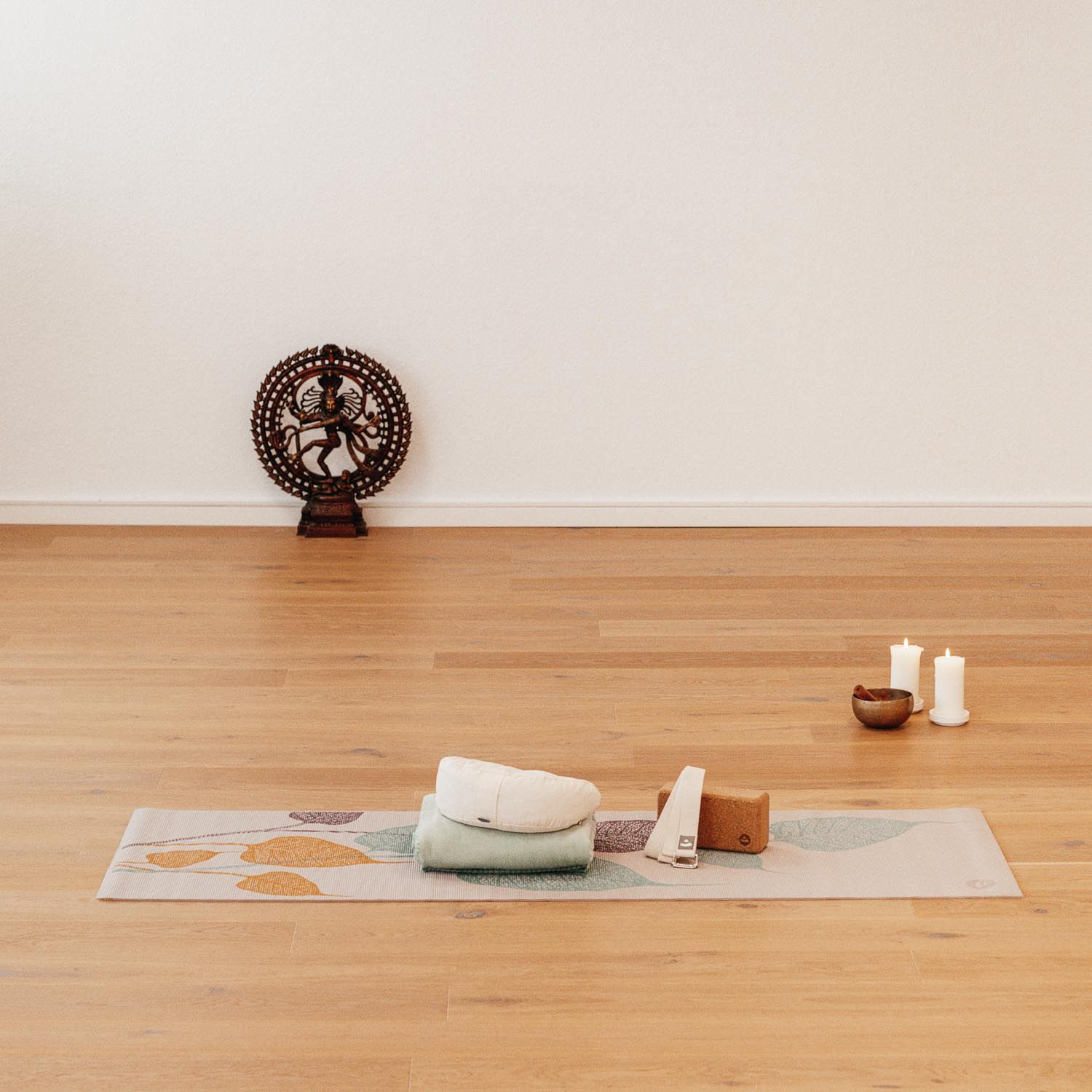 Yogamatte mit Blättermuster, beige. Auf der Matte: Handtuch, Blöcke, Kerzen, Schale. Holzfußboden, Wand im Hintergrund.