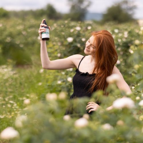 Frau hält braune Glasflasche mit Sprühkopf. Etikett mit Text: 'ALTEYA ORGANICS White Rose Water'. Frau in Blumenfeld.