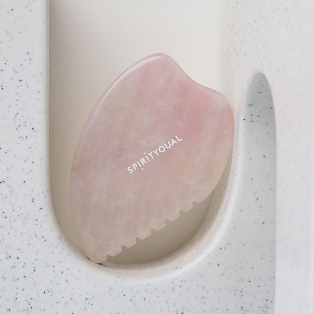 Rosenquarz Gua Sha in weißer Halterung. Rosa Stein mit abgerundeten Kanten und gezackter Seite. SPIRITYOUAL-Schriftzug.