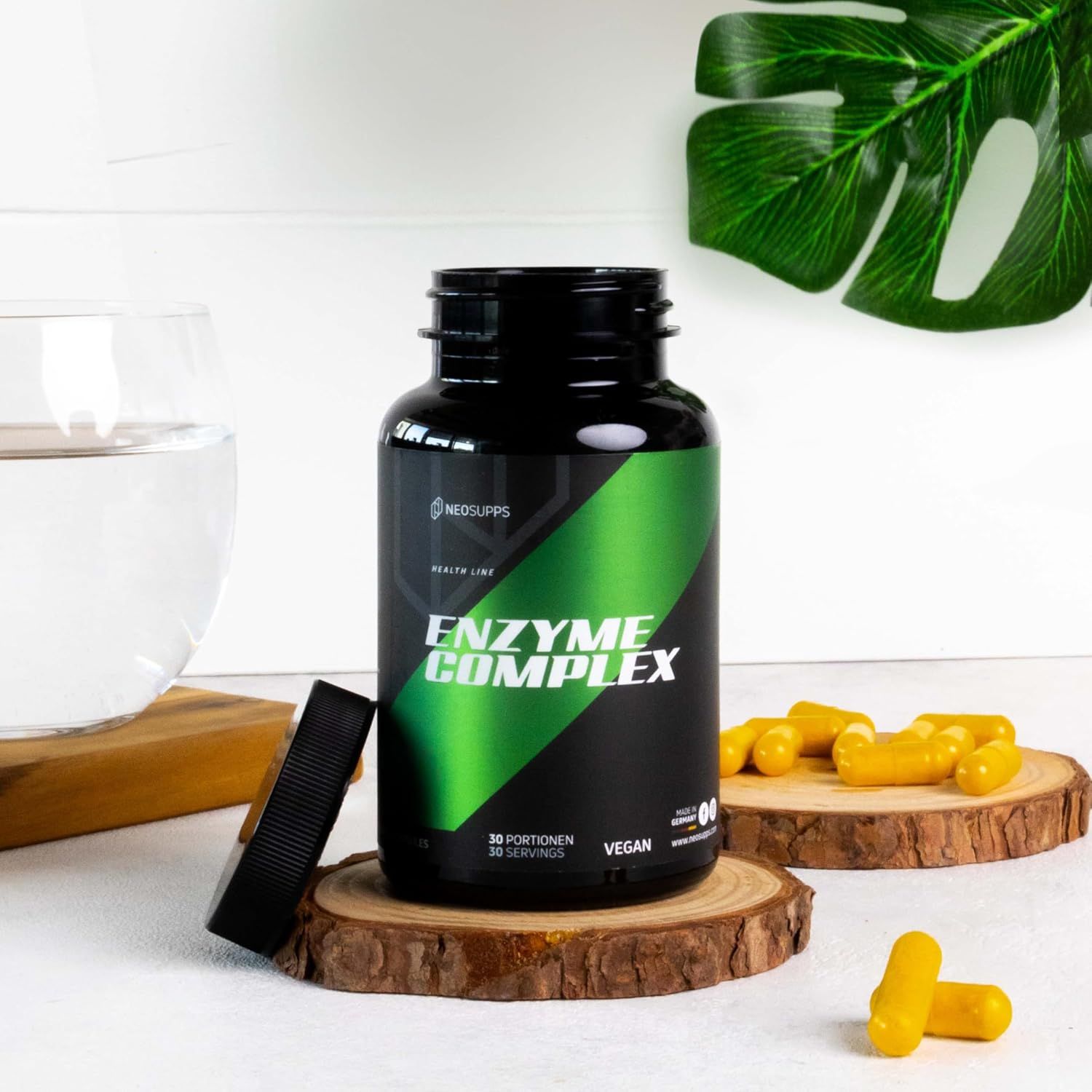Schwarze Flasche mit grünem Etikett. Text: ENZYME COMPLEX. Offener Deckel, Kapseln auf Holzscheiben. Vegan-Logo. Wasserglas.