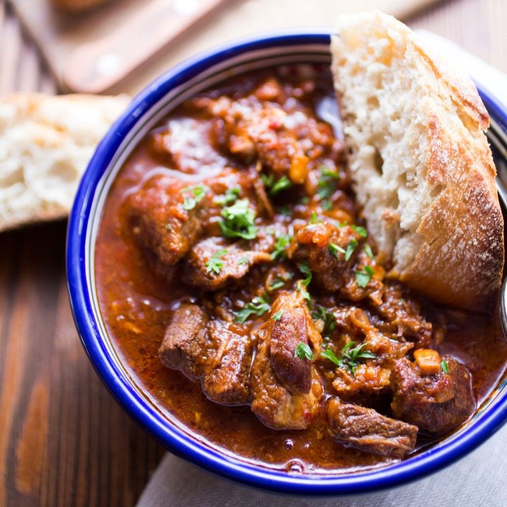 Gulasch in blauer Schüssel. Enthält Fleisch und Soße. Mit Brot serviert. Auf Holzuntergrund. Mit Petersilie garniert.