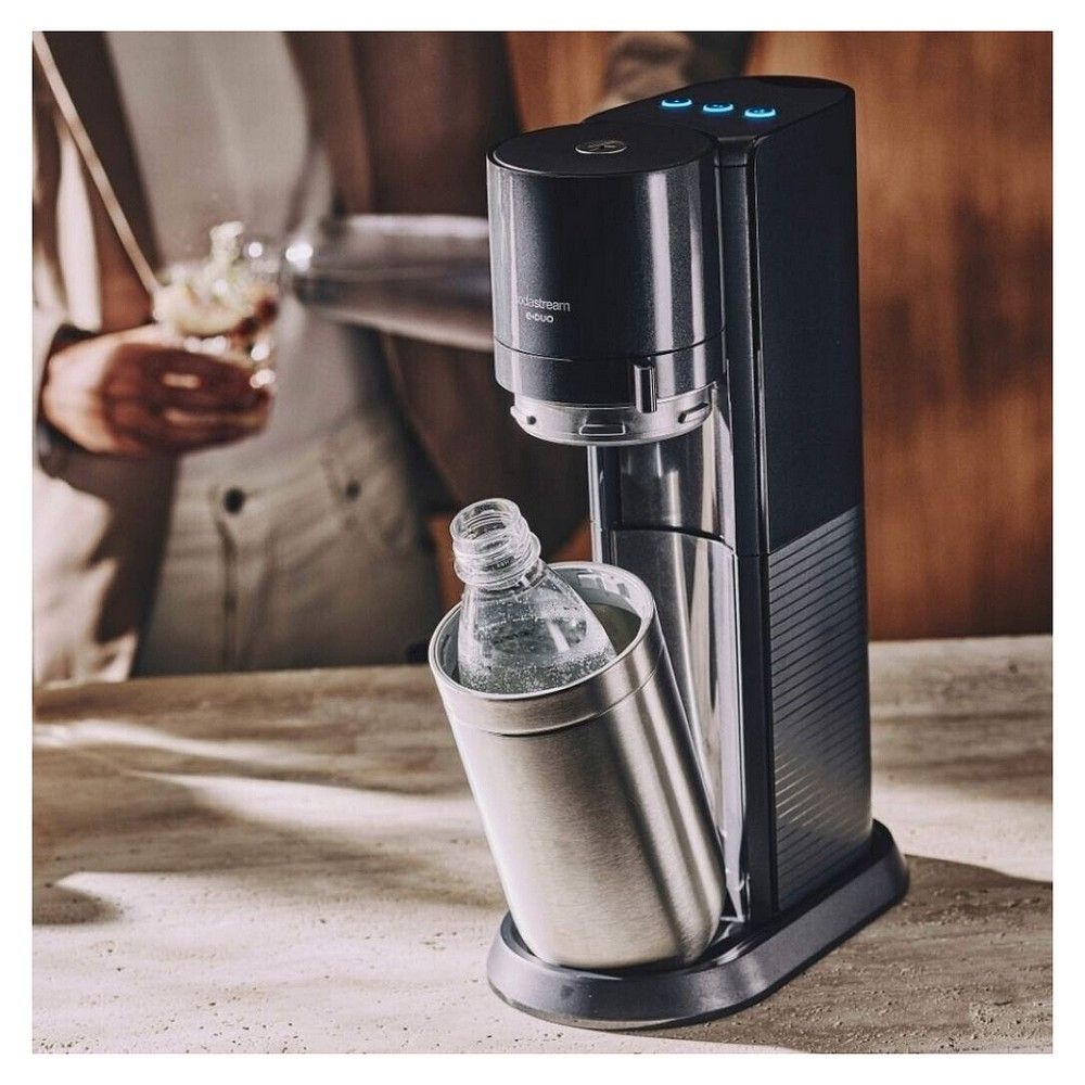 SodaStream E-DUO Wassersprudler auf einer Arbeitsfläche, im Hintergrund eine Person mit Glas.