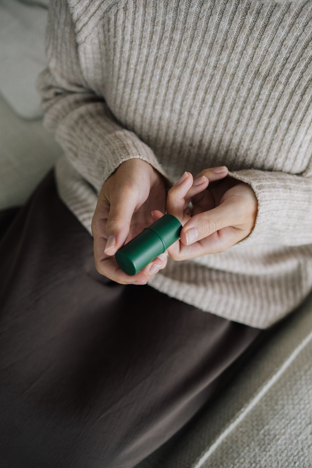 Hände halten einen grünen Lippenbalsam-Stift. Person trägt einen beigen Pullover und einen braunen Rock.