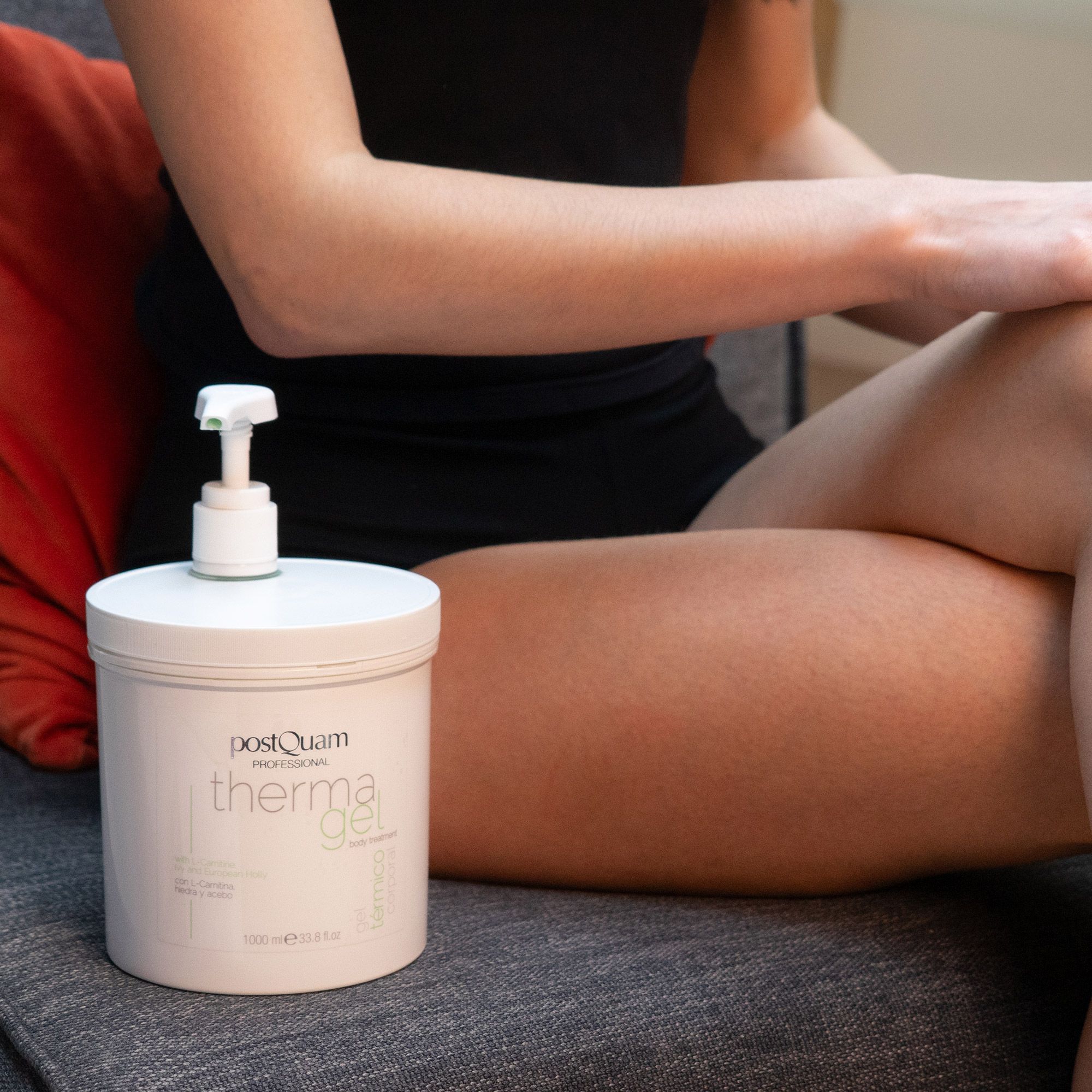 Frau sitzt mit weißem Behälter 'PostQuam therma gel'. Pumpe auf dem Behälter. Frau trägt schwarzes Oberteil.