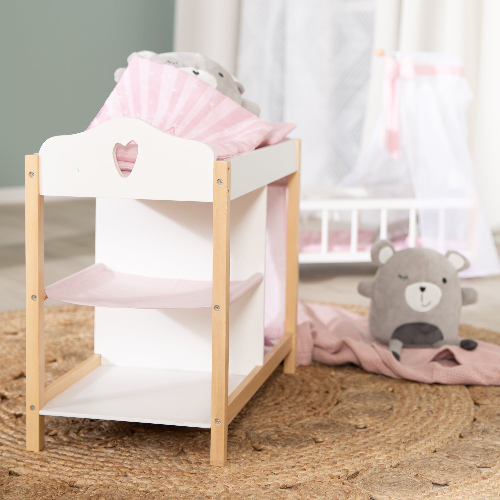 Weiße Puppenkommode mit Holzbeinen und rosa Stoffelementen. Oben ein Herz-Ausschnitt. Auf der Kommode liegen Kissen und ein Teddybär.