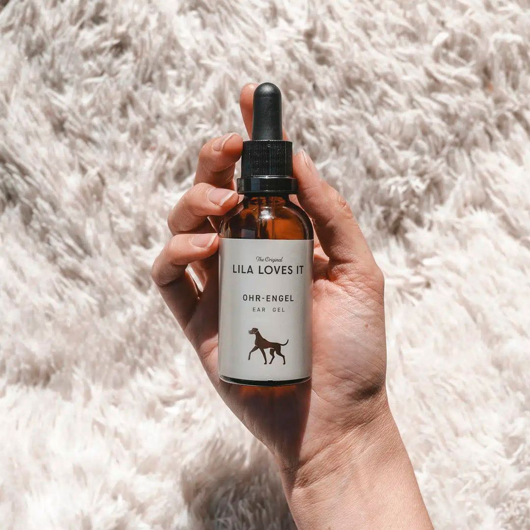 Hand hält braune Glasflasche mit Pipette. Etikett mit "LILA LOVES IT", "OHR-ENGEL" und Hund-Silhouette.