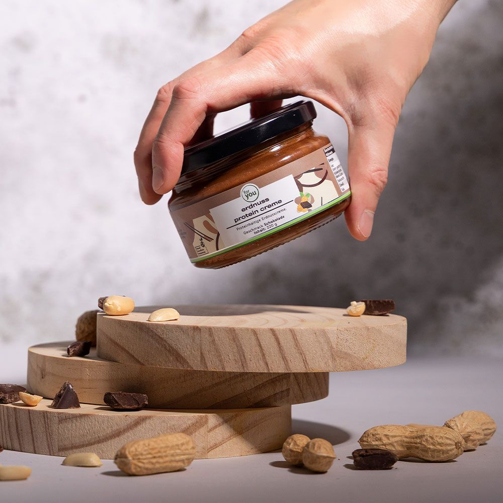 Glas mit brauner Creme, gehalten von einer Hand. Auf Holzscheiben platziert, umgeben von Erdnüssen und Schokoladenstücken.