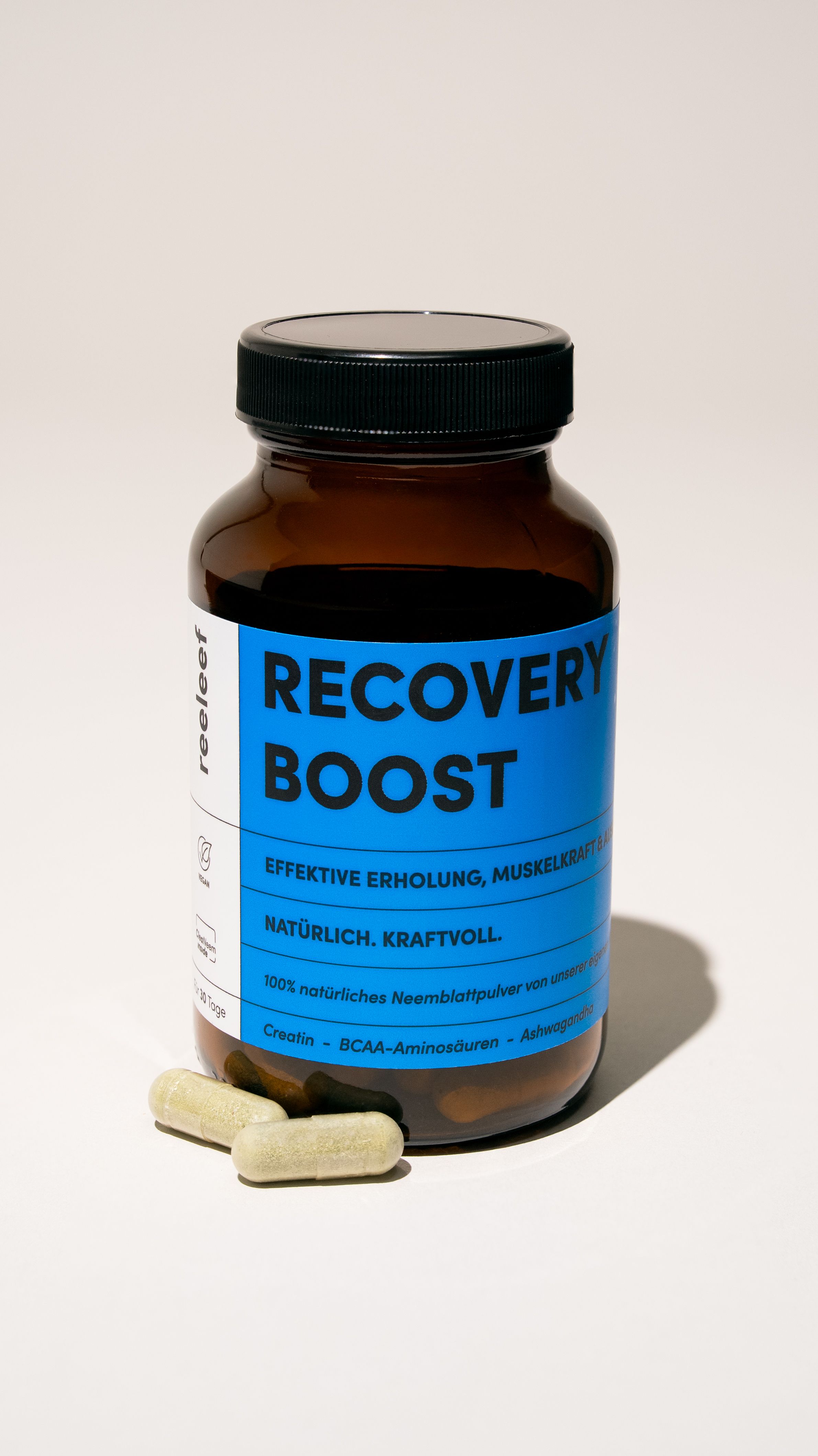 Braune Glasflasche mit schwarzem Deckel und blauem Etikett. Aufschrift: Recovery Boost. Daneben liegen drei Kapseln.