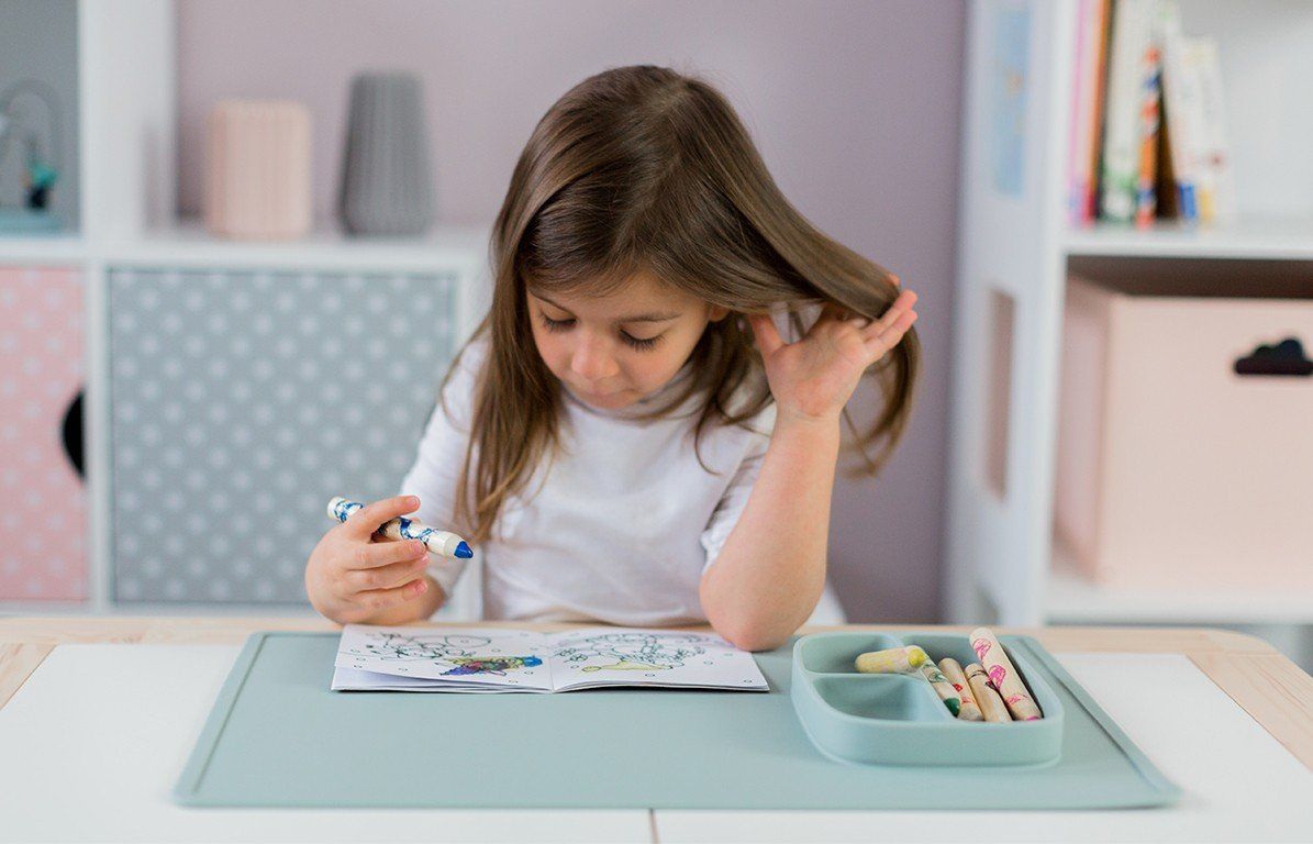 Kind sitzt an Tisch, malt mit Stift. Aquamarinfarbene Spielmatte mit integriertem Teller. Buntstifte im Teller.