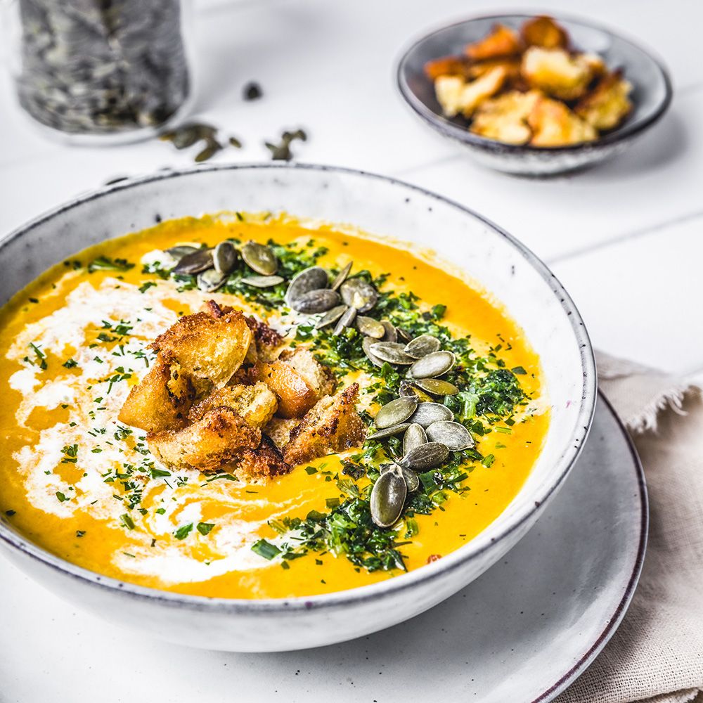 Schüssel mit Kürbissuppe, Sahne, Croutons, Kürbiskernen und Kräutern. Auf Teller.