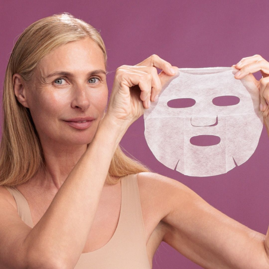 Frau hält Gesichtsmaske vor violettem Hintergrund. Maske mit Augen-, Nasen- und Mundöffnungen. Helle Haut, blondes Haar.