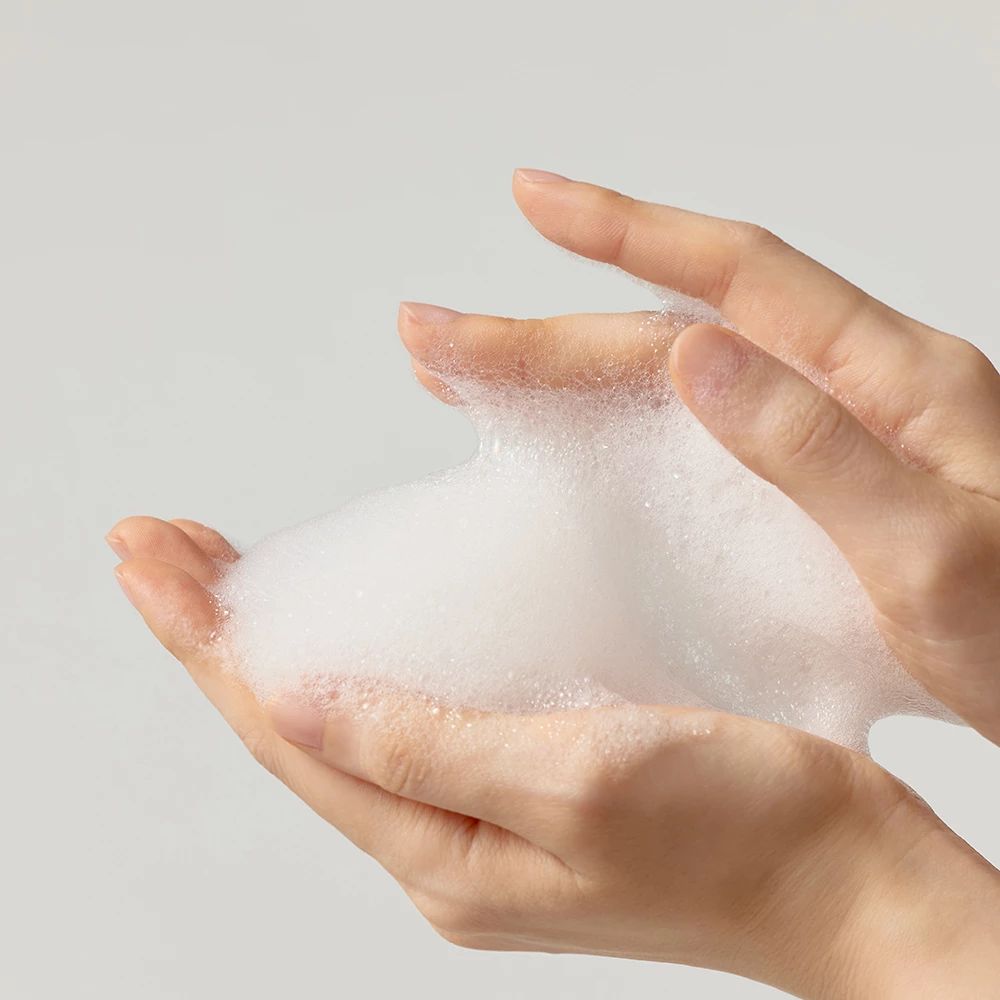 Hände halten weißen Schaum. Der Schaum ist dicht und luftig. Helle Haut. Hintergrund ist hellgrau.