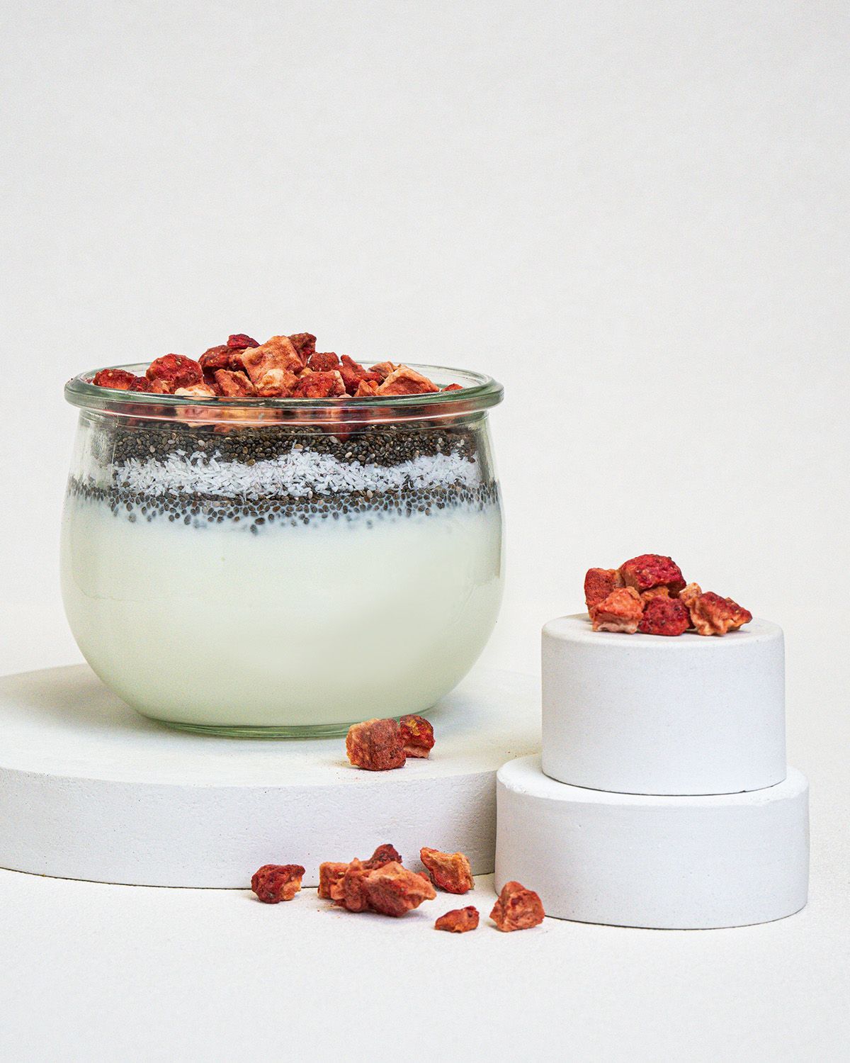 Glas mit Joghurt, Chia-Samen und gefriergetrockneten Erdbeerstücken. Erdbeerstücke auf Podesten.