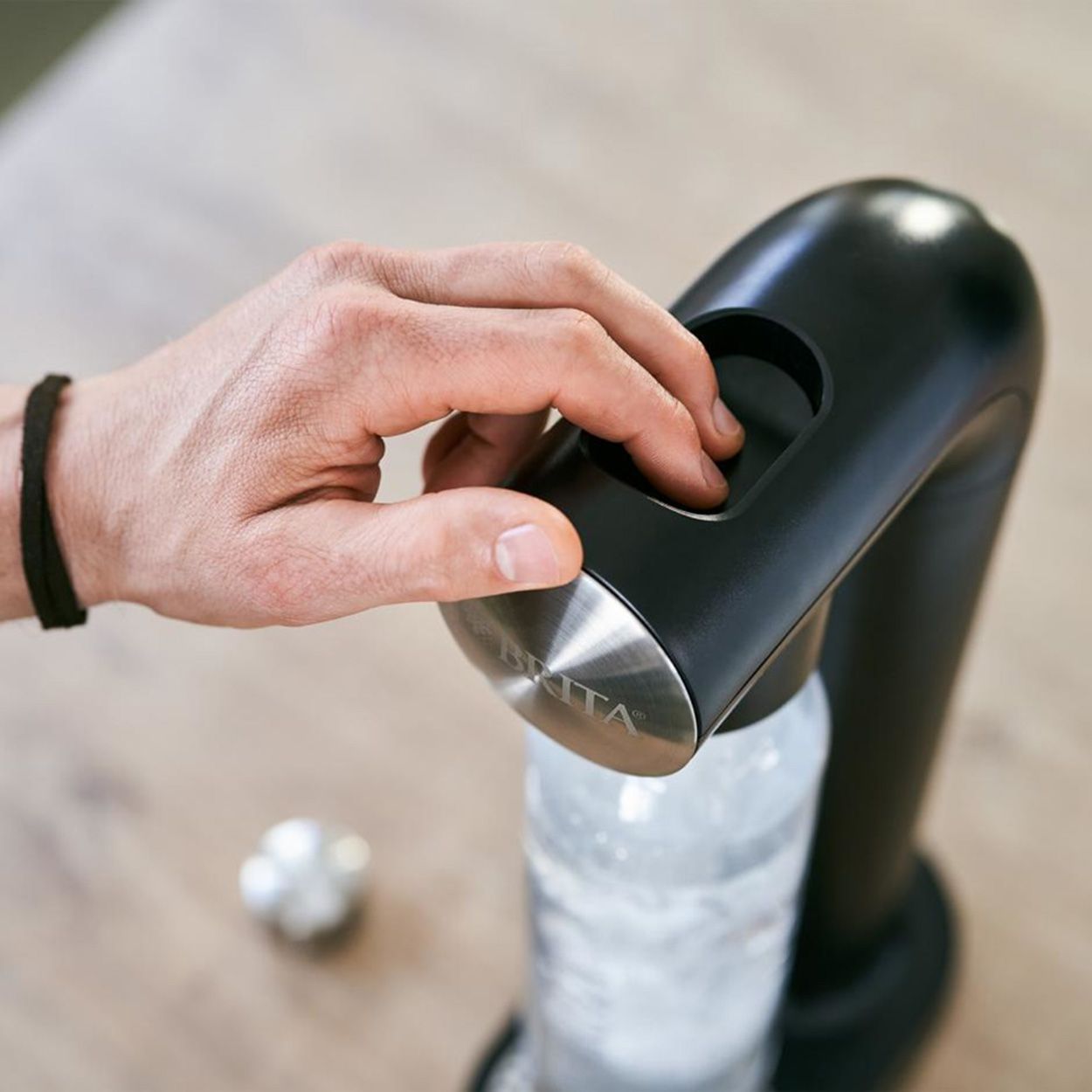Der BRITA sodaONE Wassersprudler in Schwarz wird von einer Hand bedient. Der Sprudler hat eine silberne Oberfläche und eine transparente Flasche. Auf dem Tisch liegt ein silberner Knopf.