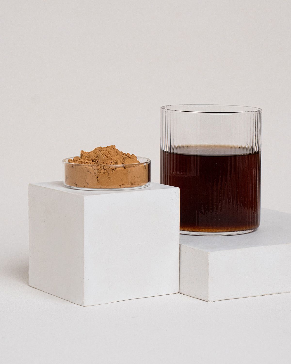 Glas mit Flüssigkeit und Schale mit braunem Pulver auf weißen Blöcken. Hintergrund hell.