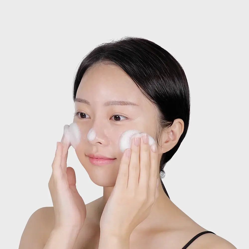 Frau wäscht ihr Gesicht mit Schaum. Helle Haut, dunkle Haare. Weißer Hintergrund.