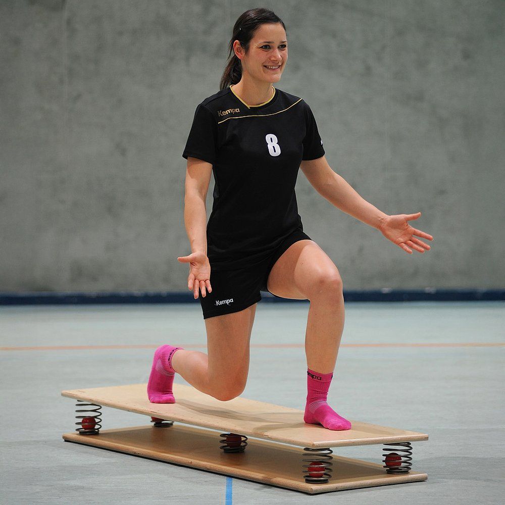 Person auf Federbrett, 150 x 45 cm, in Ausfallschritt-Position. Schwarze Sportkleidung.