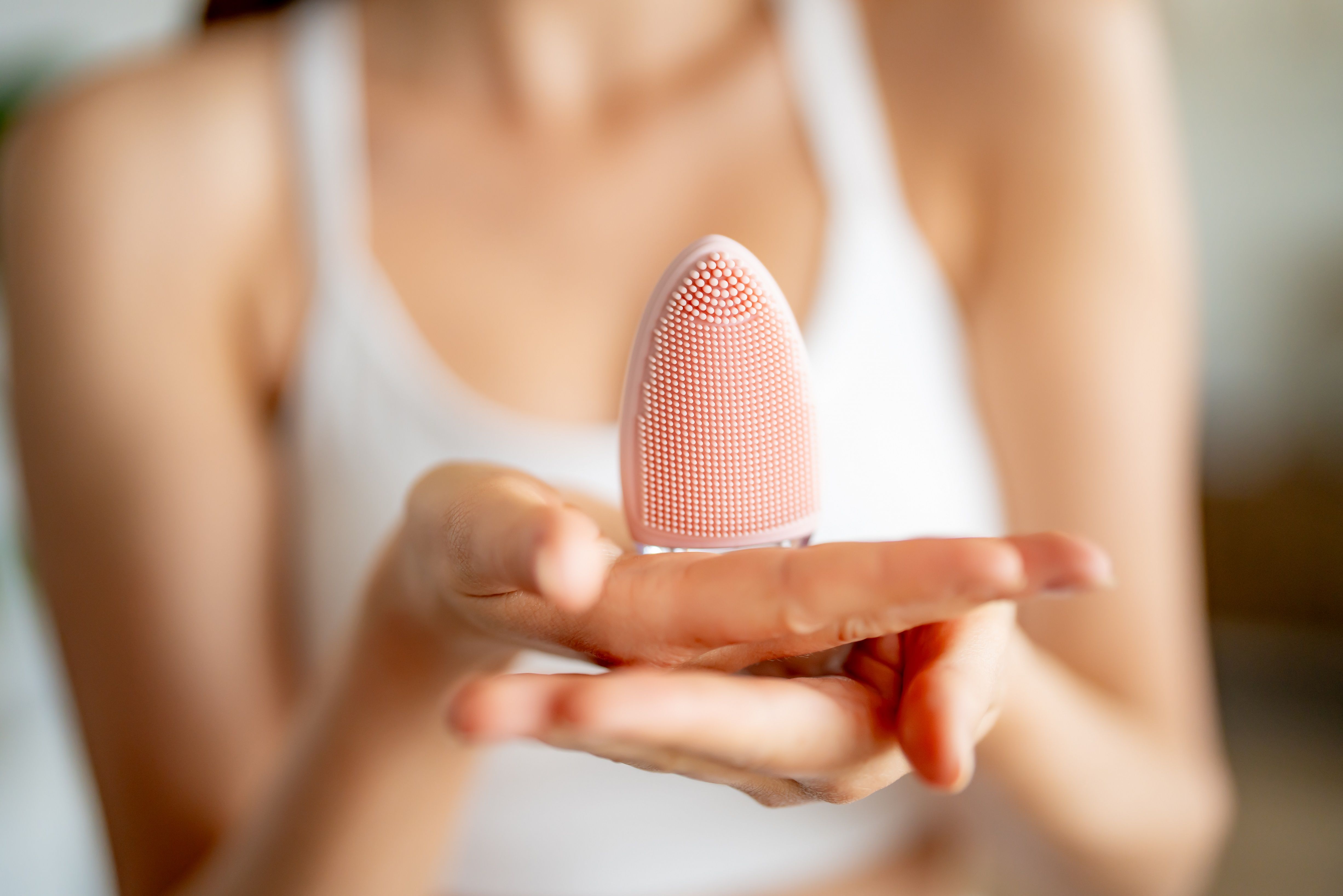 Hände halten rosa Schall-Gesichtsbürste. Ovaler Bürstenkopf mit Silikonborsten. Helle Umgebung.