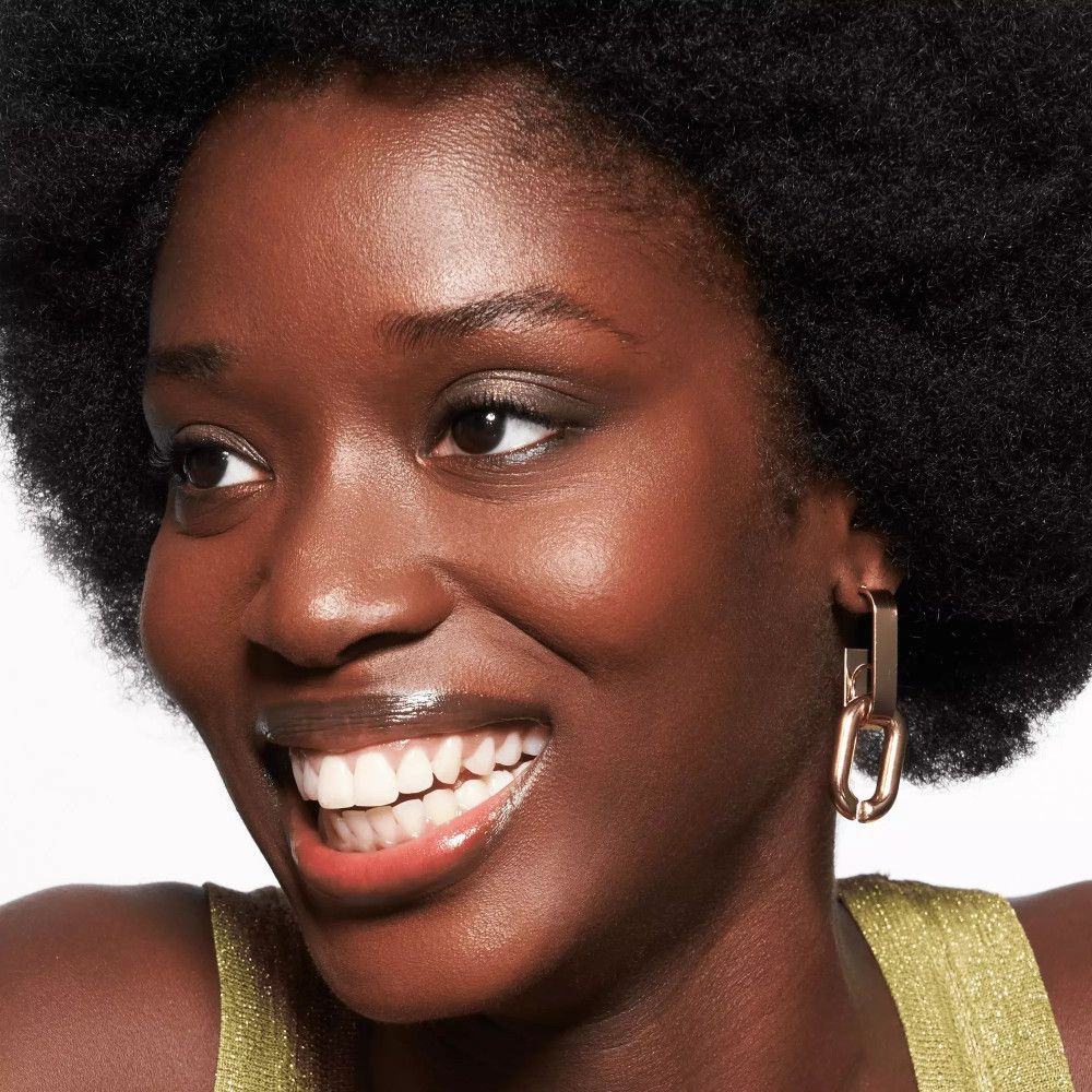 Porträt einer lächelnden Frau mit dunkler Haut und Locken. Sie trägt goldene Ohrringe und glänzenden Lippenbalsam.