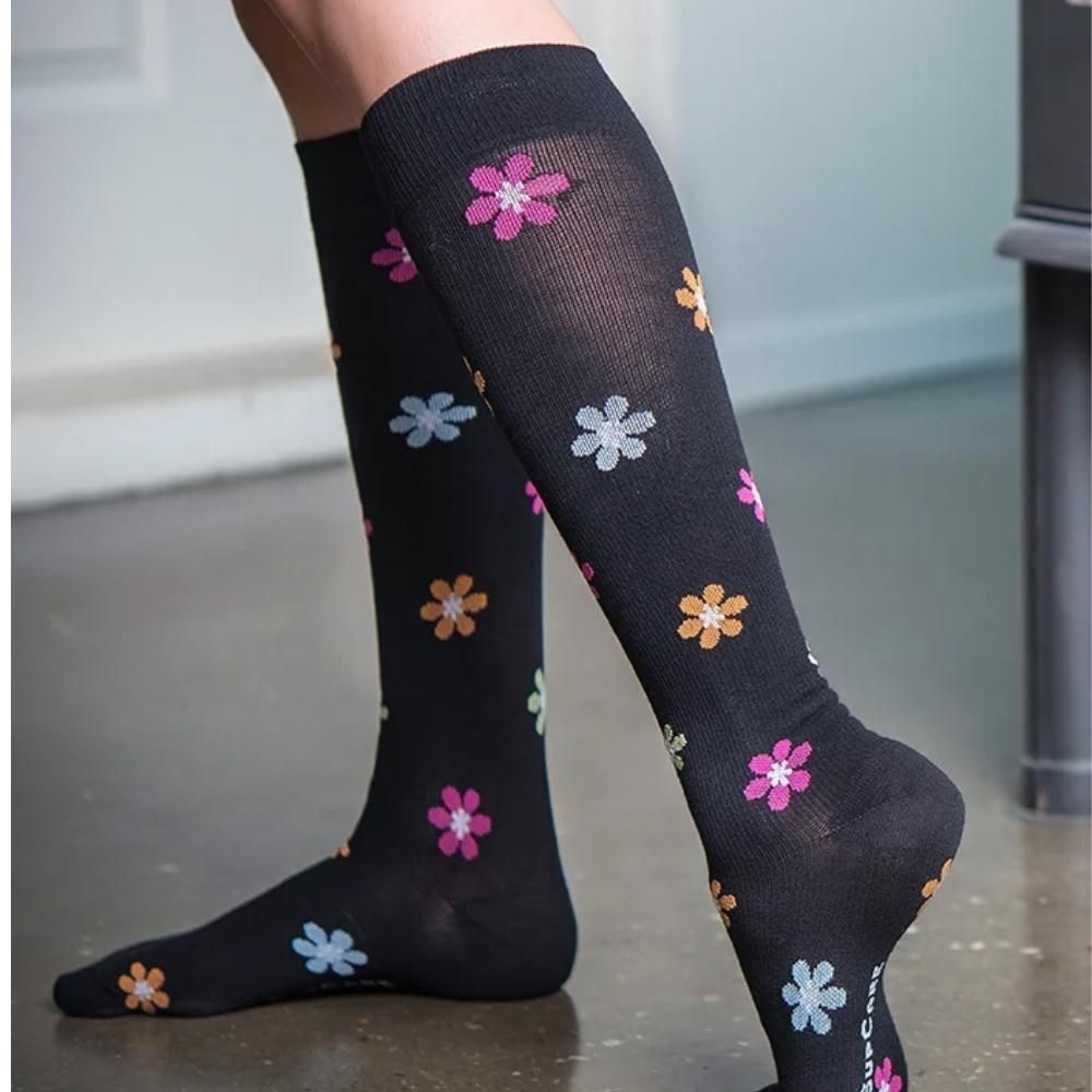Person trägt schwarze Stützstrümpfe mit bunten Blumen. Marke SupCare am Fuß. Kniestrümpfe.