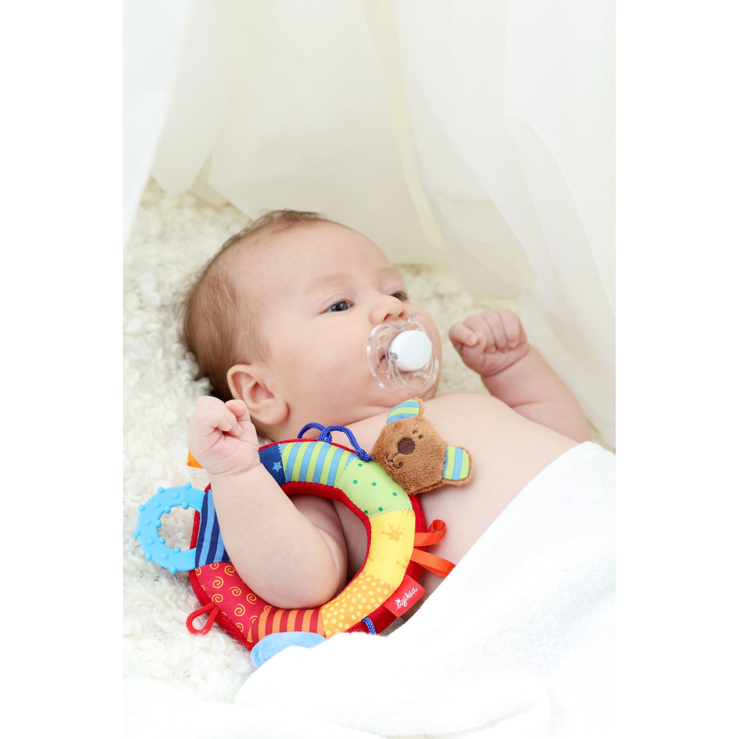 Baby liegt mit Schnuller und Ringgreifling auf einer weißen Decke. Der Ringgreifling ist sichtbar.