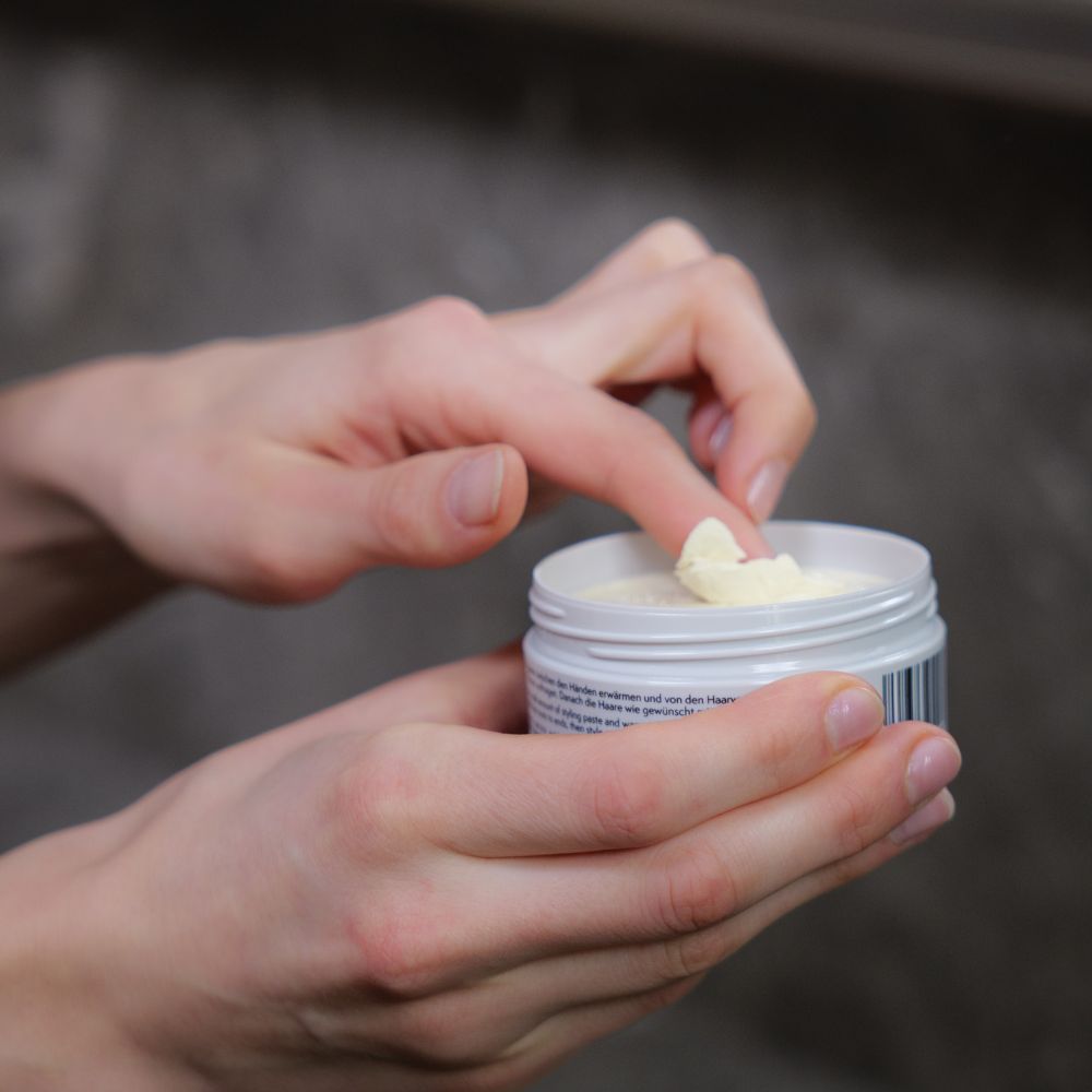 Hände öffnen Genki Styling Paste Dose. Grauer Deckel, schwarzer Schriftzug, gelber Farbfleck. Creme.