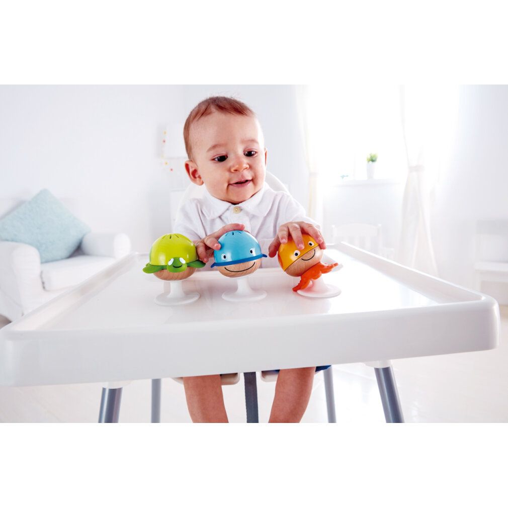 Baby sitzt an einem Tisch und spielt mit drei Rassel-Spielzeugen. Blaues Wal-Design, gelbes Tintenfisch-Design und grünes Schildkröten-Design.