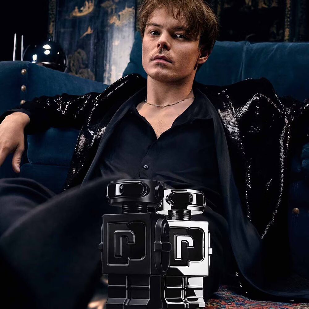 Mann sitzt auf Sofa, hält zwei Roboterflakons. Einer silber, einer schwarz. Schwarze Kleidung.