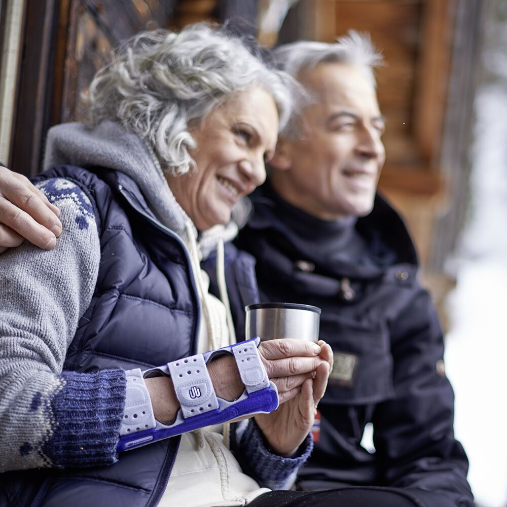 Handgelenk-Daumenorthese ManuLoc Rhizo, blau-grau, am Handgelenk einer Person. Im Hintergrund ein älteres Ehepaar.