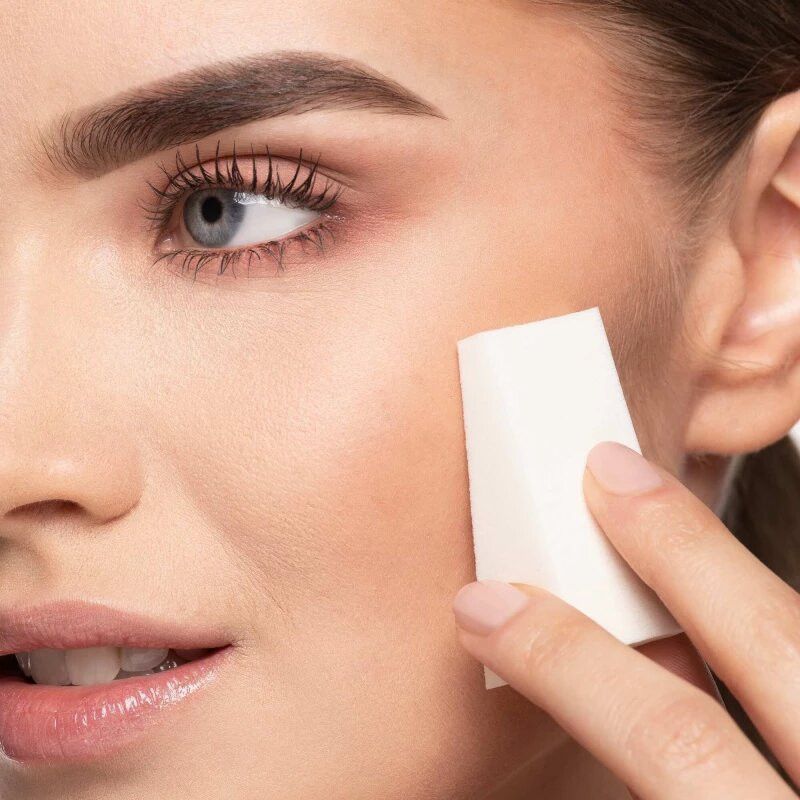 Eine Frau trägt mit einem weißen Make-up-Schwamm Foundation auf ihre Wange auf.
