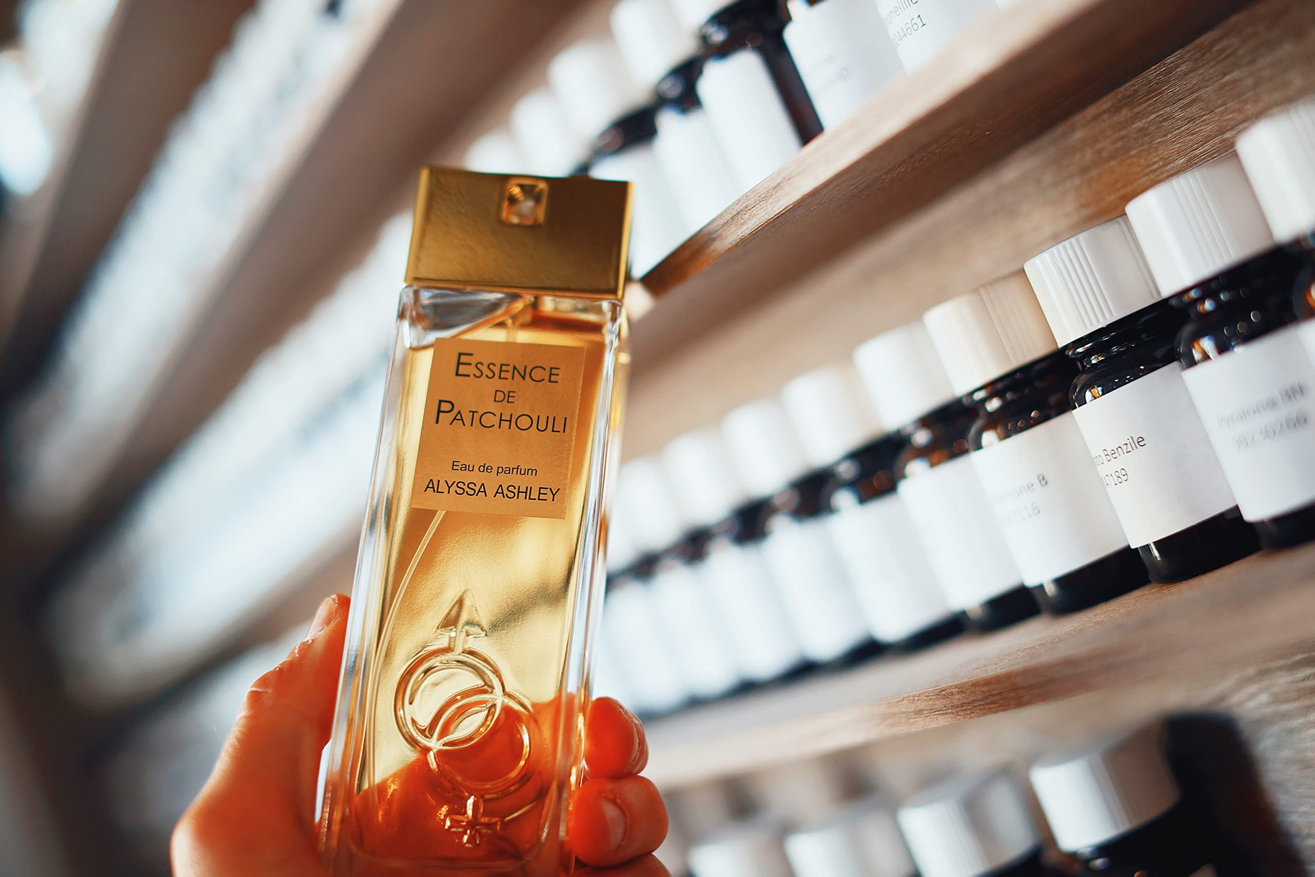 Person hält Flakon. Aufschrift: Essence de Patchouli, Eau de parfum, Alyssa Ashley. Goldener Flakon mit Sprühkopf. Regale im Hintergrund.