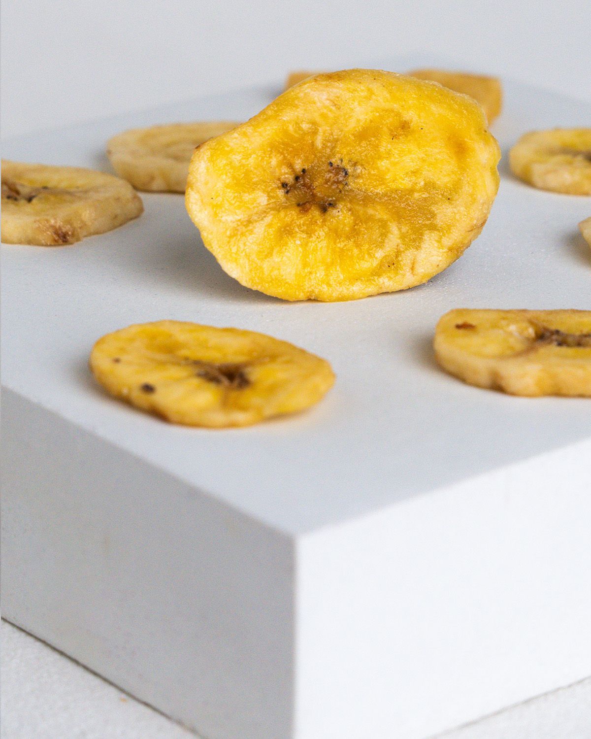 Mehrere Bio-Bananenchips auf einer weißen Oberfläche. Einzelne Chips, gelbliche Farbe, leicht gebräunt. Auf einem weißen Podest.