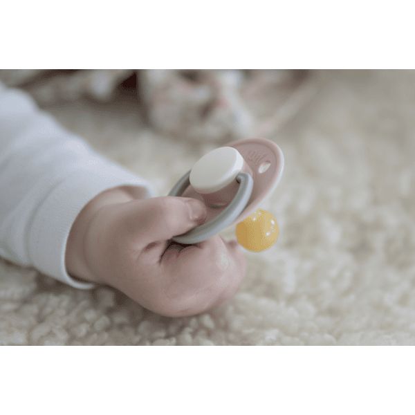 Babyhand hält Schnuller. Der Schnuller hat einen weißen Sauger und einen grauen Rand.