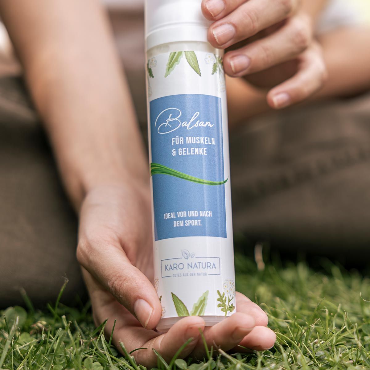Produkt in Händen gehalten. Zylindrische Flasche mit blauem Etikett. Text 'Balsam für Muskeln & Gelenke'. Karo Natura Logo.