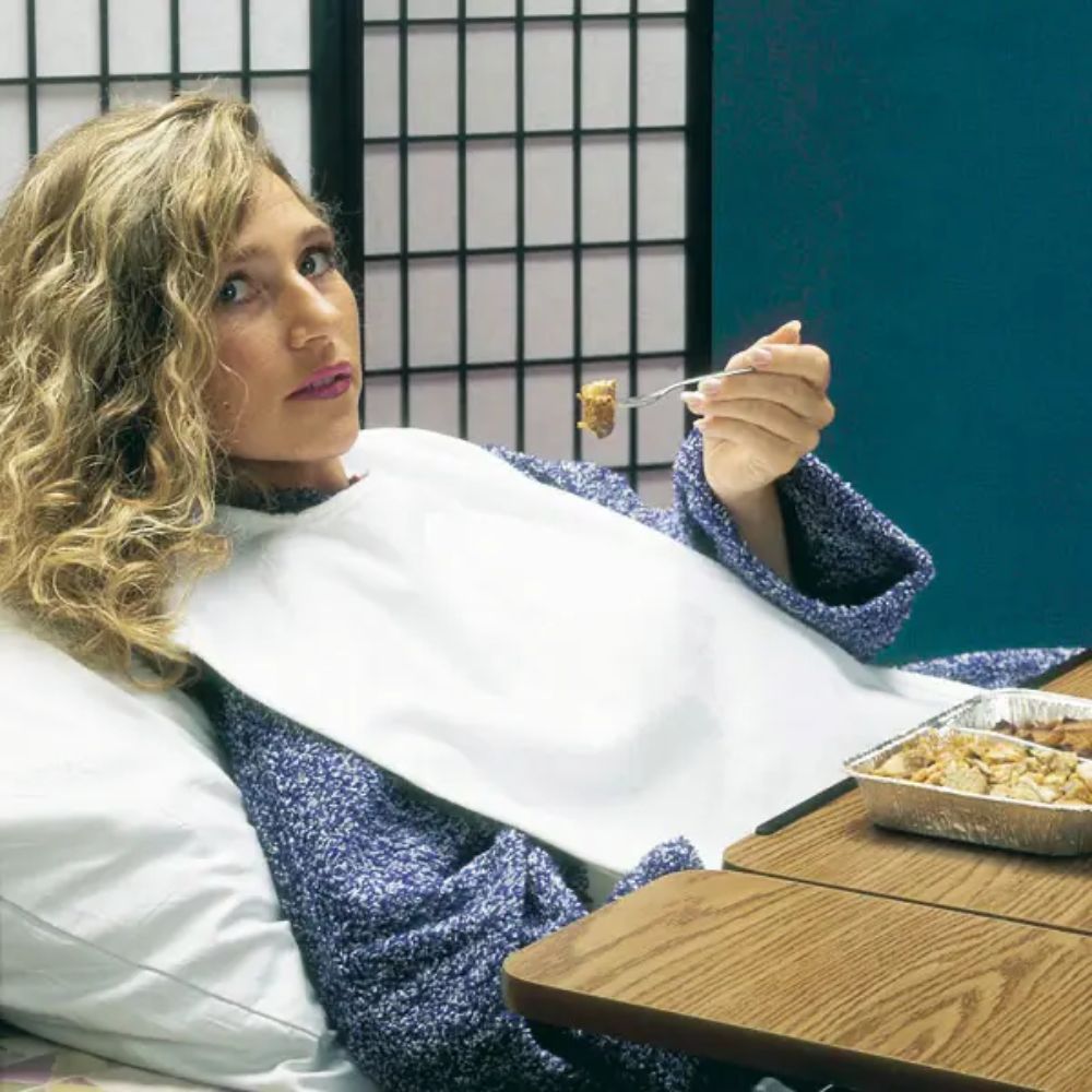 Weißer Kleiderschutz für Erwachsene. Die Person trägt den Schutz beim Essen. Auf einem Tisch stehen Essensbehälter.