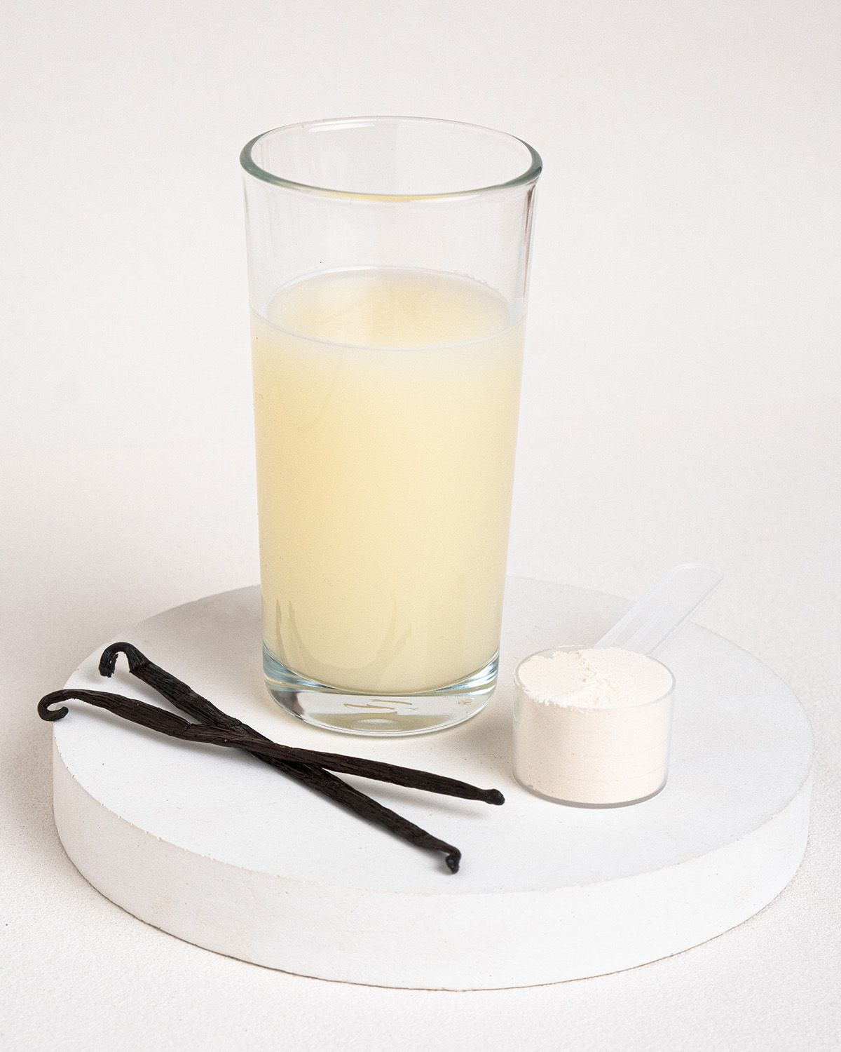 Glas mit Flüssigkeit, Löffel mit Pulver, Vanilleschoten auf weißem Podest. Helles Getränk, weißes Pulver, braune Schoten.